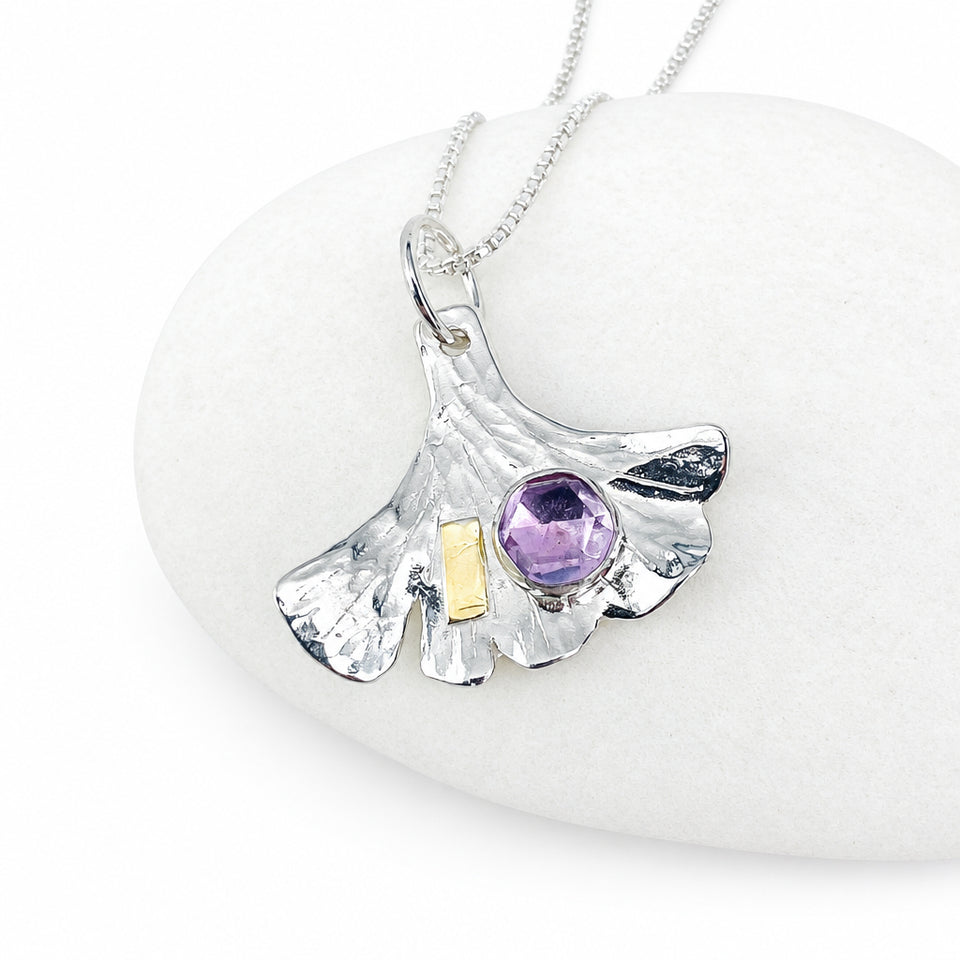 A silver pendant necklace shaped like a ginkgo leaf, featuring a faceted purple gemstone and a small gold accent, displayed on a smooth white stone.
