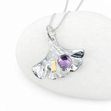 A silver pendant necklace shaped like a ginkgo leaf, featuring a faceted purple gemstone and a small gold accent, displayed on a smooth white stone.