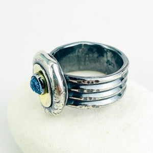 A handmade silver ring with a textured, multi-band design and a central oval gold-toned inlay featuring a round blue topaz gemstone, displayed on a white surface.