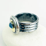 A handmade silver ring with a textured, multi-band design and a central oval gold-toned inlay featuring a round blue topaz gemstone, displayed on a white surface.