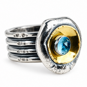 A textured sterling silver ring with a multi-band wrap design, featuring a gold circular inlay and a central round-cut faceted blue topaz gemstone.