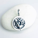 A sterling silver circular pendant with an embossed bird flying free from prison bars motif design, with light patina for contrast, resting on a smooth white stone against a light background.