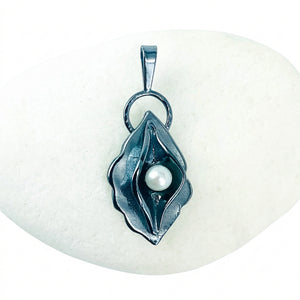 A dark metallic pendant shaped like a stylized flower or leaf with a single white pearl in the center, displayed on a smooth white stone against a light background.