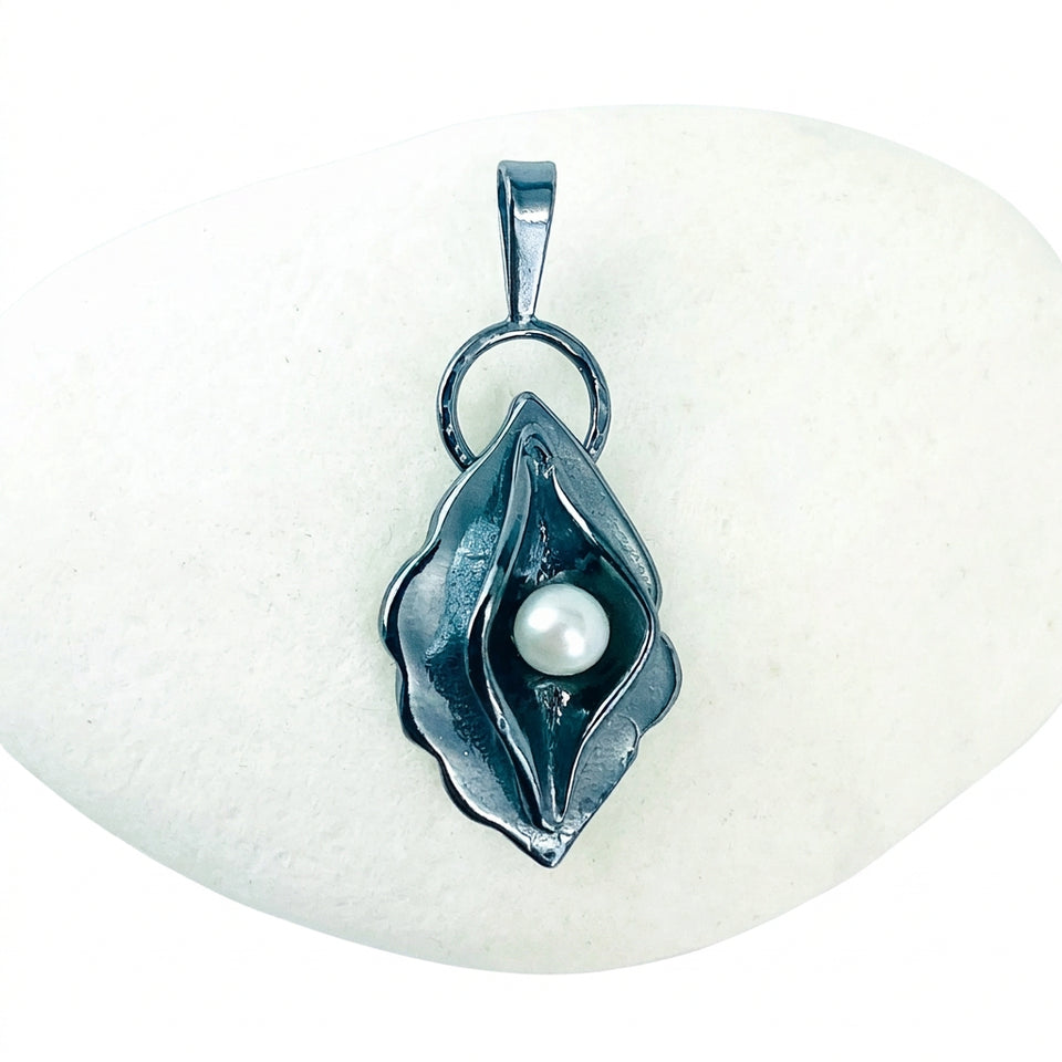 A dark metallic pendant shaped like a stylized flower or leaf with a single white pearl in the center, displayed on a smooth white stone against a light background.