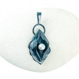 A dark metallic pendant shaped like a stylized flower or leaf with a single white pearl in the center, displayed on a smooth white stone against a light background.