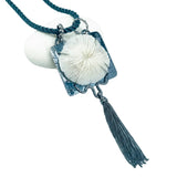 A gunmetal silver-toned pendant necklace featuring a textured square frame, a large white mushroom coral in the center, and a long gunmetal silver tassel, displayed on a white background with a small white stone prop.