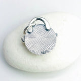 A sterling silver round back of pendant with a textured wavy pattern, featuring '925' marking, resting on a smooth white stone against a white background.