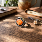 A silver ring with a large oval orange spiny oyster stone, resting on a rustic wooden table with books, a pen, and dried flowers in the background.