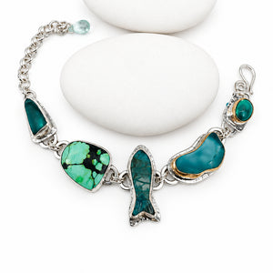 A sterling silver chain bracelet with multiple green and turquoise, chrysoprase and sea glass charms, displayed on a white background with three stacked white stones.