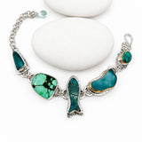 A sterling silver chain bracelet with multiple green and turquoise, chrysoprase and sea glass charms, displayed on a white background with three stacked white stones.