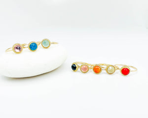 gold stackable with round multicolor gemstone settings, one displayed on a white stone and the other on a white surface, showcasing a range of gemstone colors including purple, blue, bluish green, garnet, pink, orange, and red.