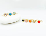 gold stackable with round multicolor gemstone settings, one displayed on a white stone and the other on a white surface, showcasing a range of gemstone colors including purple, blue, bluish green, garnet, pink, orange, and red.