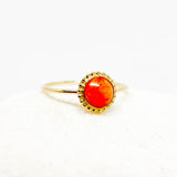 A gold ring with a round, vibrant reddish orange ruby cabochon gemstone and a beaded gold halo, displayed on a soft white textured surface.