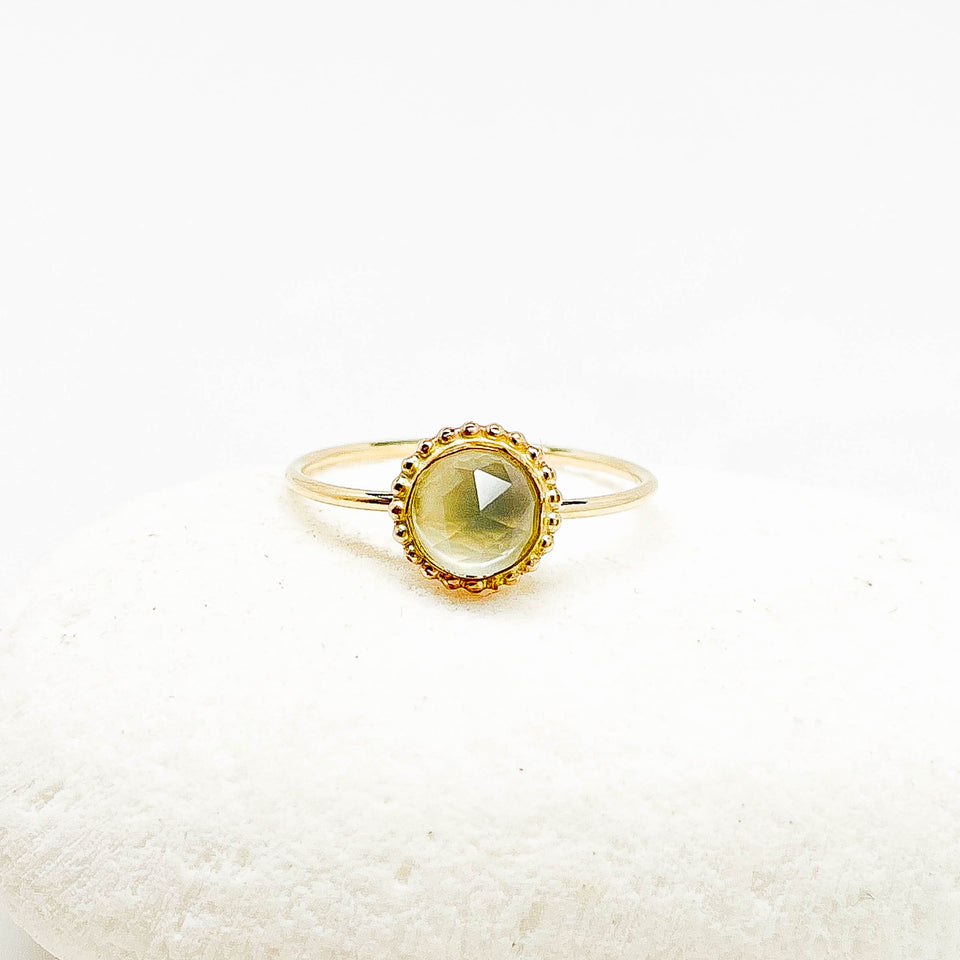 A gold ring with a round faceted light moonstone gemstone, displayed on a white textured stone against a white background.