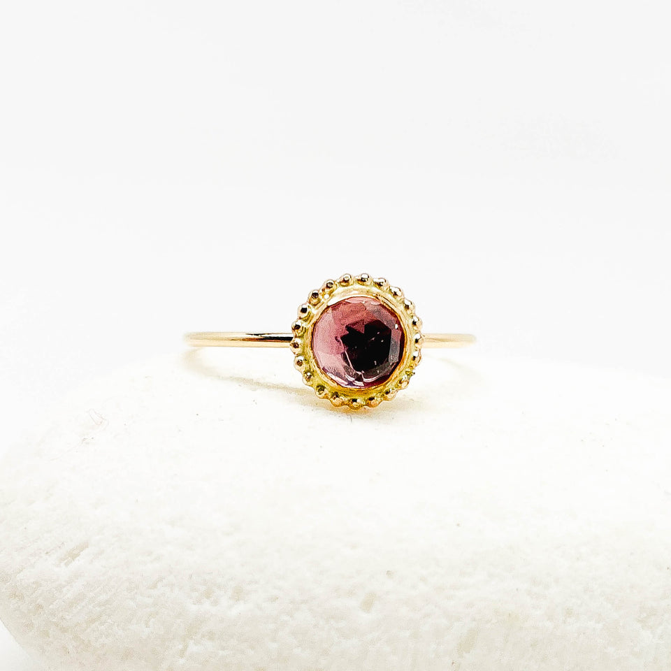 A gold ring with a round faceted amethyst gemstone and a decorative beaded gold border, displayed on a textured white stone against a white background.