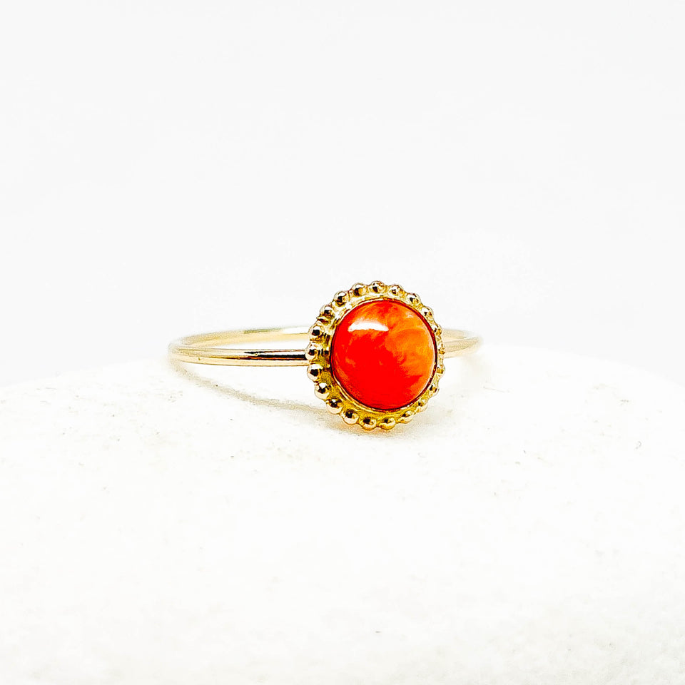 A gold ring with a round, vibrant orange cabochon gemstone, set in a scalloped gold bezel on a white textured background.