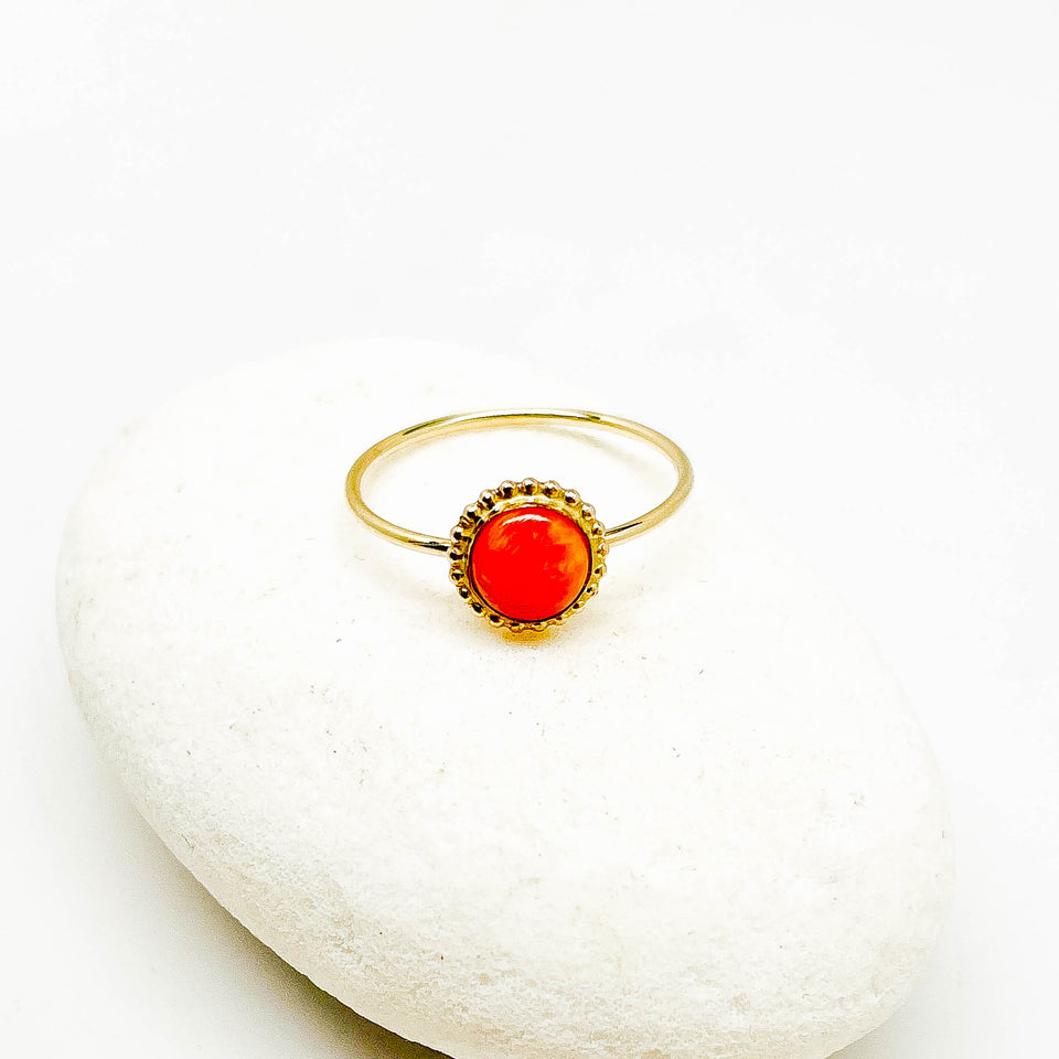 A gold ring with a round, vibrant orange-red ruby cabochon gemstone, displayed on a smooth white stone against a white background.