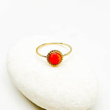 A gold ring with a round, vibrant orange-red ruby cabochon gemstone, displayed on a smooth white stone against a white background.