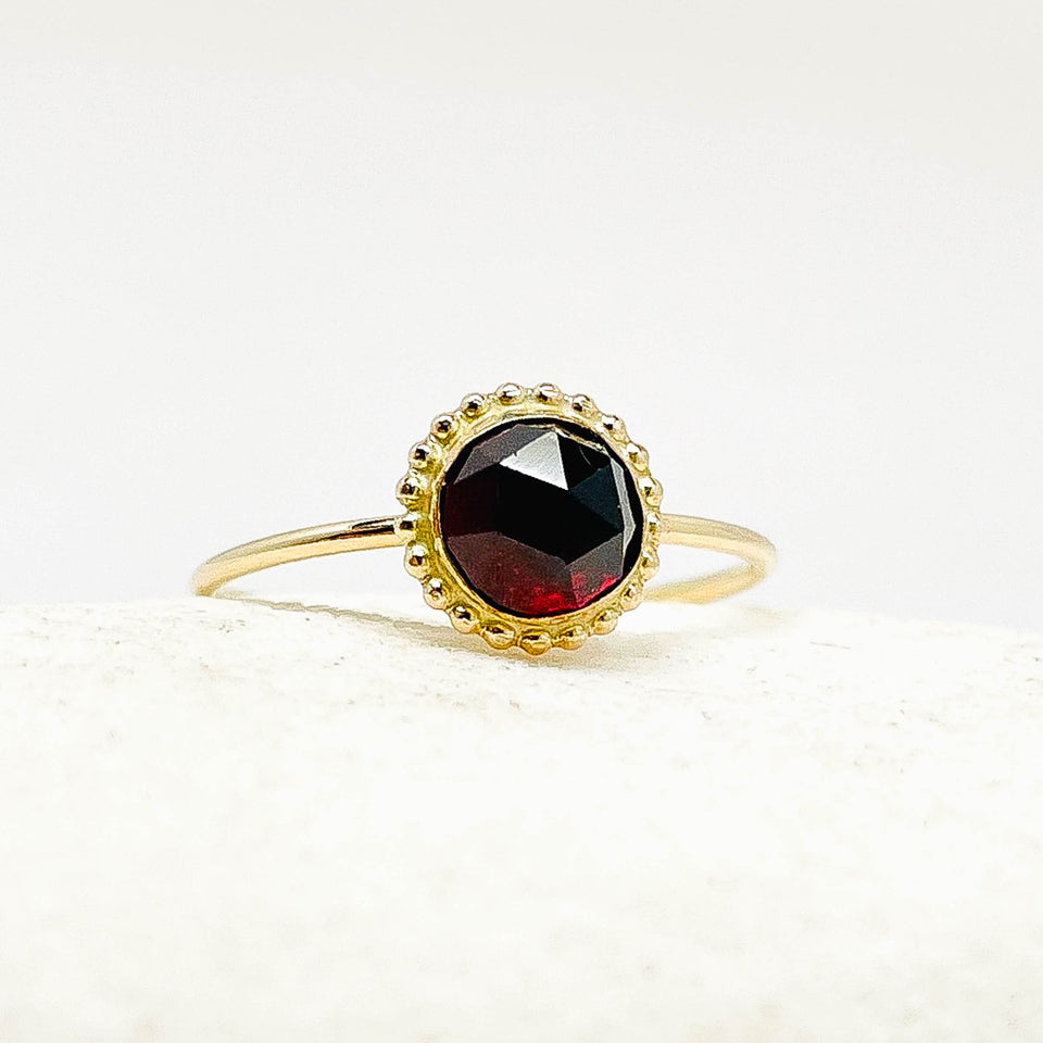 A gold ring with a round faceted dark red garnet gemstone, set in a scalloped gold bezel and resting on a textured white surface.