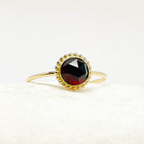 A gold ring with a round faceted dark red garnet gemstone, set in a scalloped gold bezel and resting on a textured white surface.