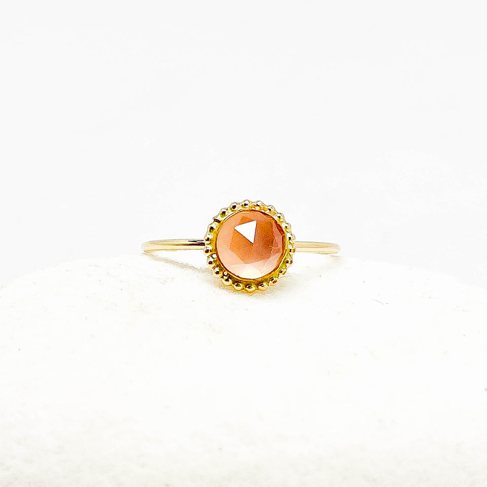 A gold ring with a round faceted rose quartz gemstone, displayed on a soft white textured surface.