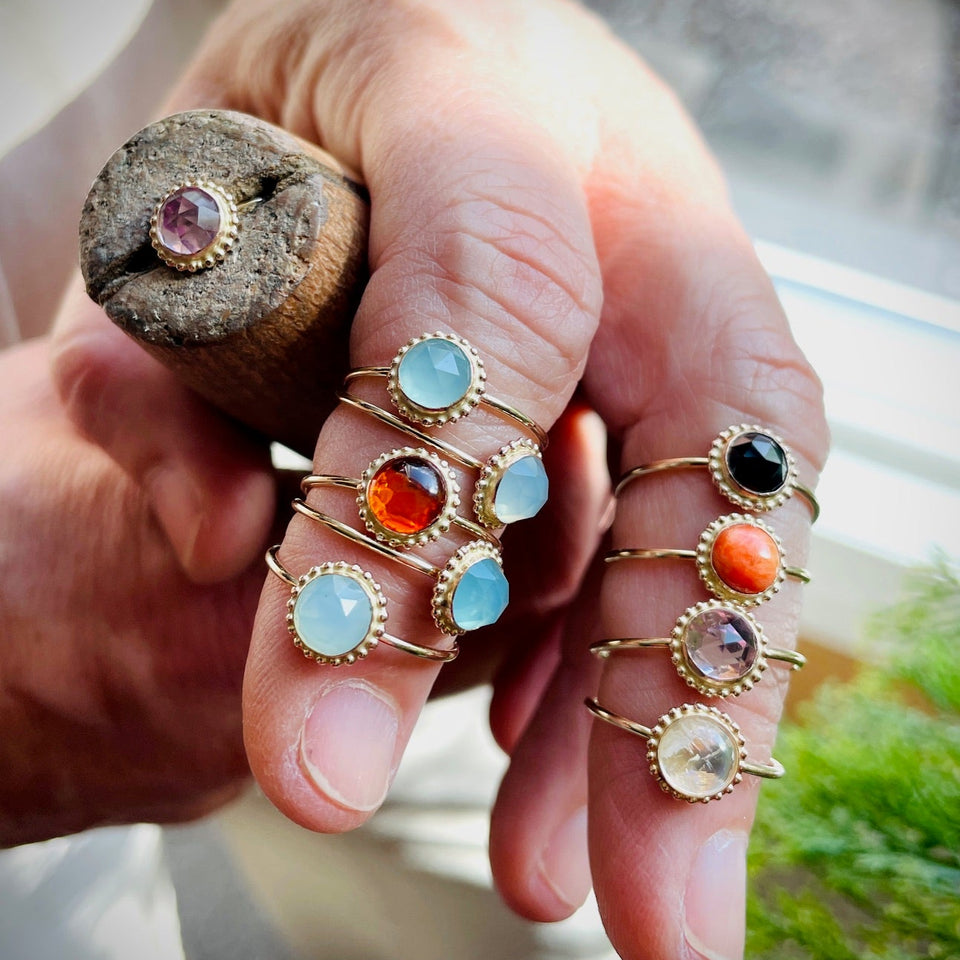 A close-up of a hand wearing multiple gold rings with round gemstones in light blue, orange, black, purple, and clear settings, while holding a rustic wooden block with a pink gemstone.